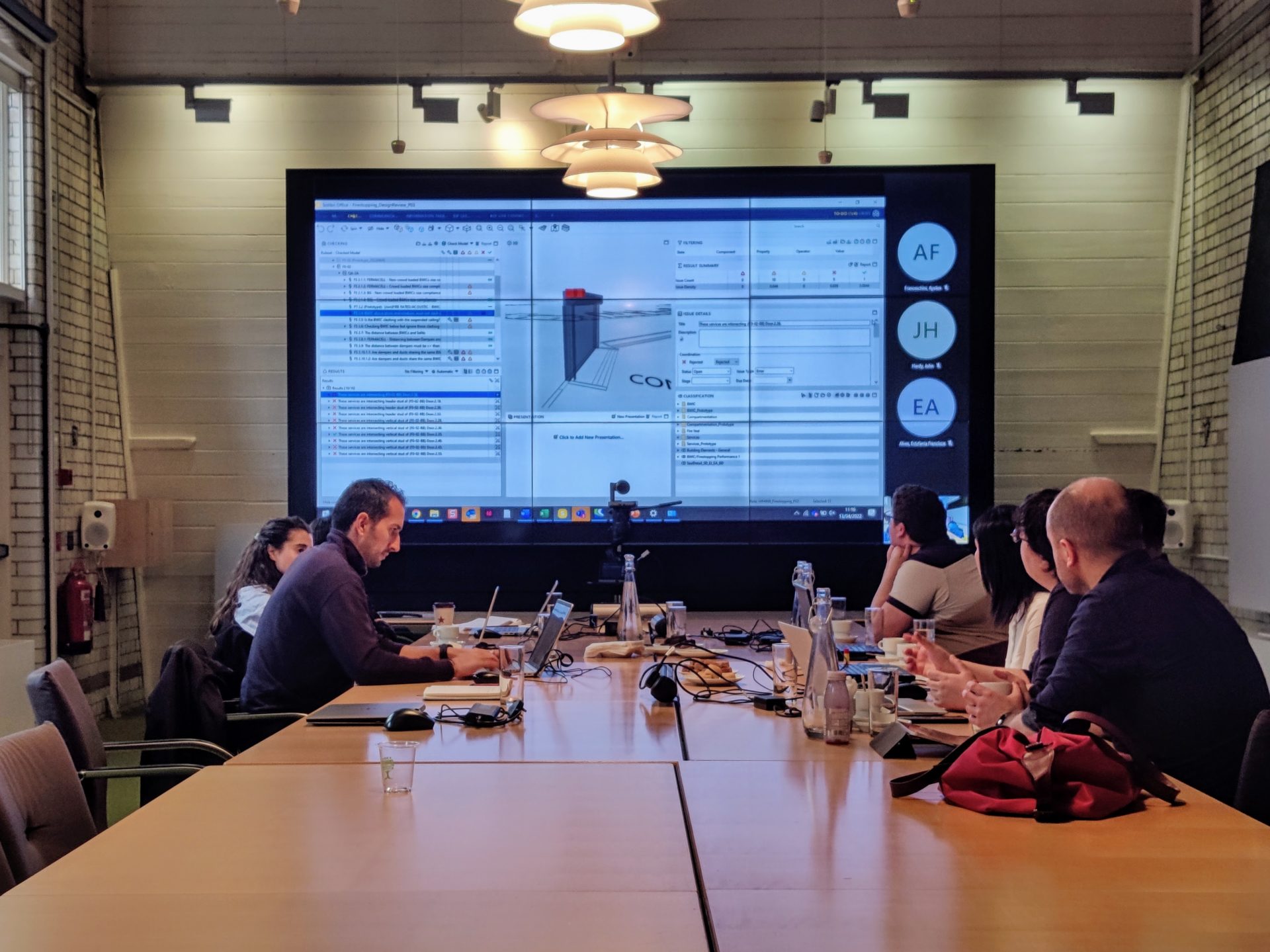 People seating around a big table with laptops looking at a big screen showing data in it. 