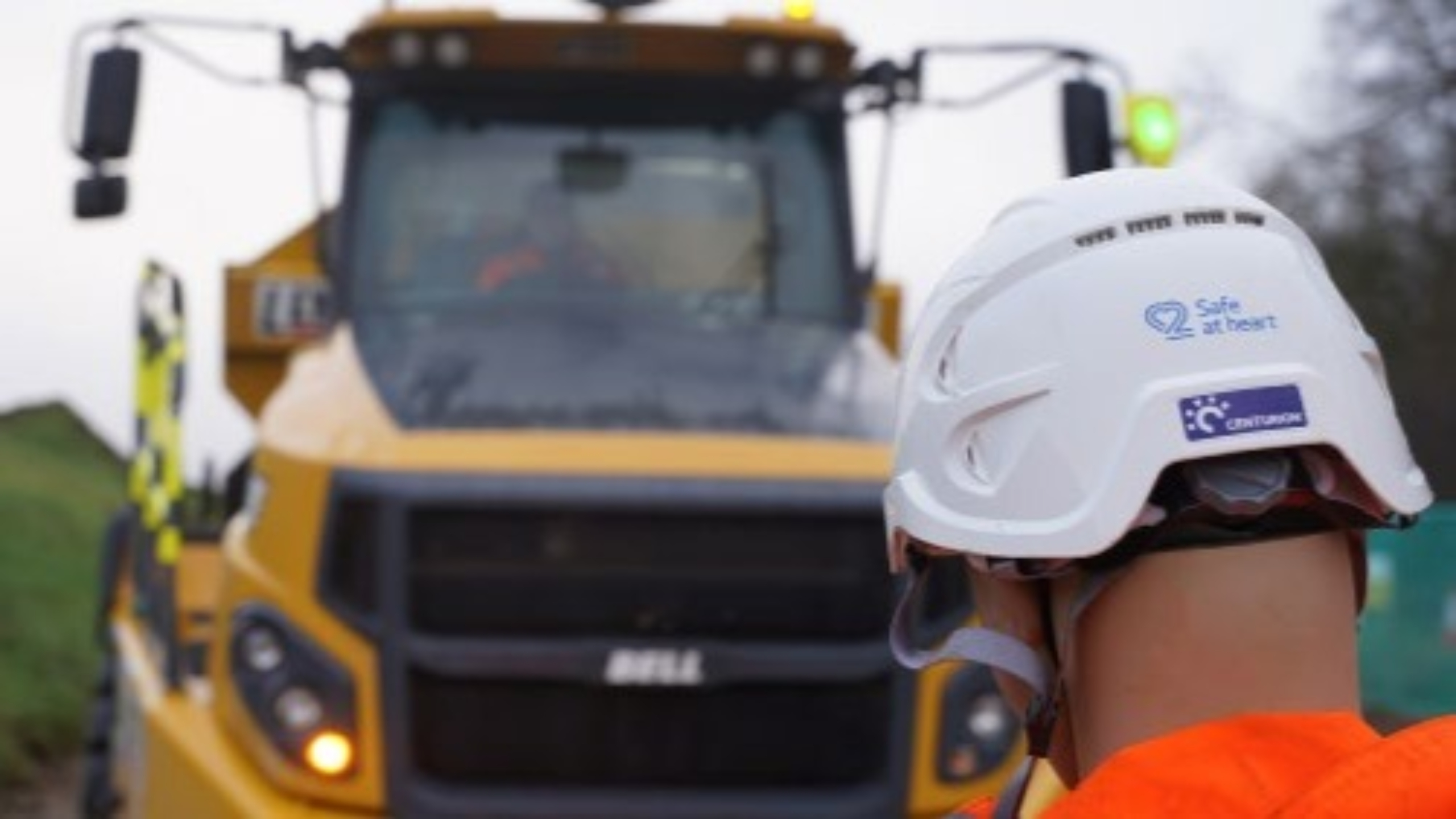 A yellow construction vehicle in front of a person wearing a white hard helmet.